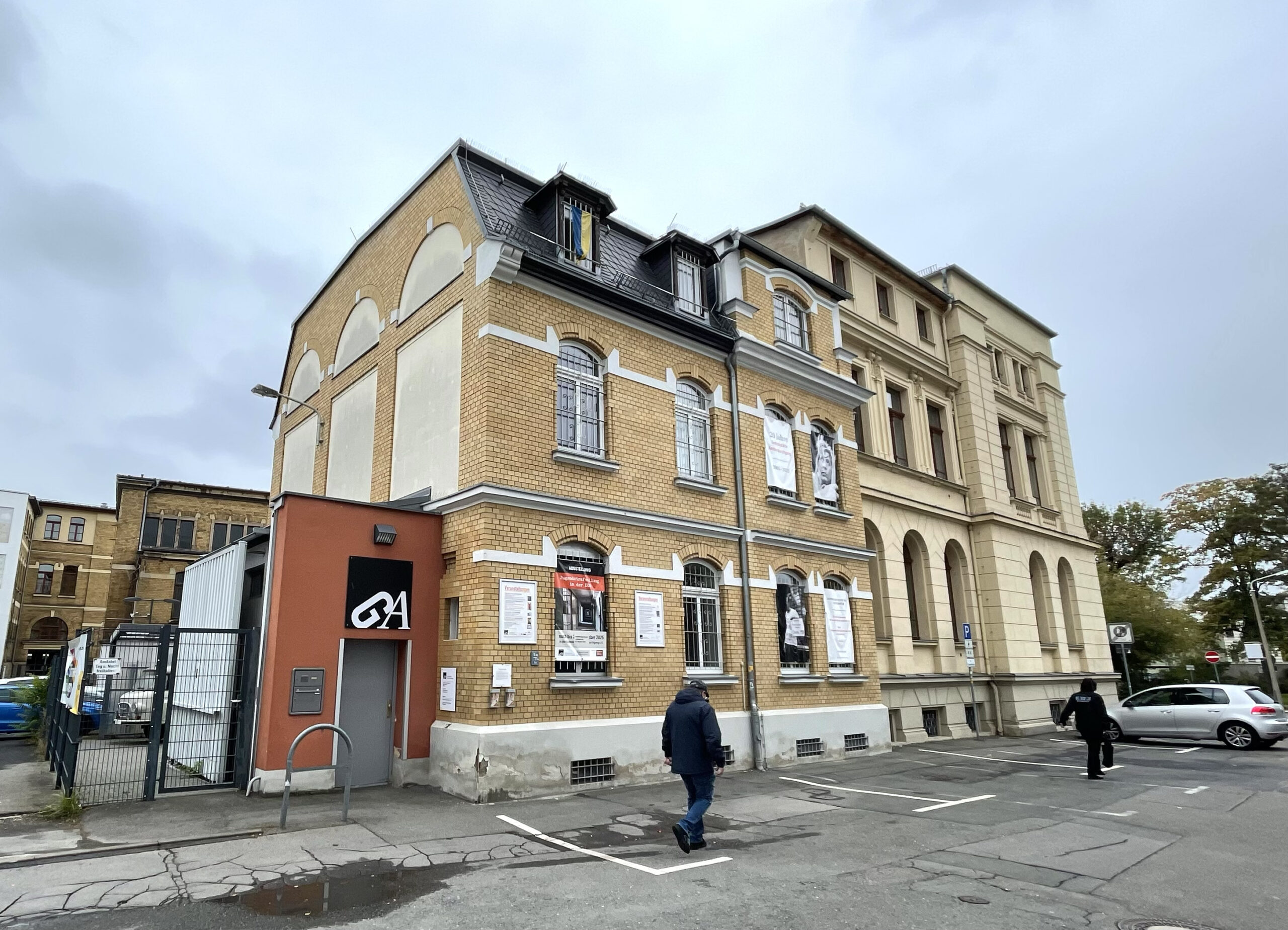 Backsteingebäude mit mansardenförmigem Dach, orange Anbau mit Logo, zwei Personen auf Gehweg, historisches Ensemble an Straße
